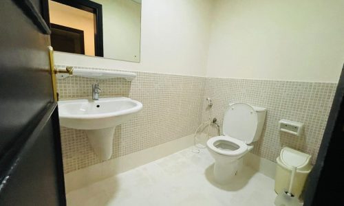 A small bathroom in a furnished apartment for rent, with a white toilet, bidet, and pedestal sink, featuring beige tiled walls and a large mirror.