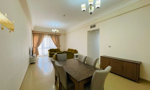 Spacious living room in a furnished apartment with a dining table, sofa, and large windows with curtains. Neutral color palette with tiled flooring in the Sanabis area.