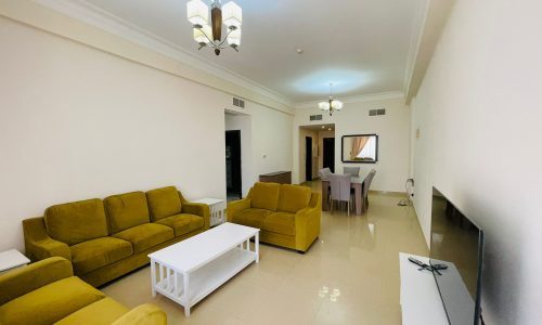 A spacious living room in a furnished apartment with mustard yellow sofas, a white coffee table, a flat-screen tv, and an adjoining dining area with a table and chairs.