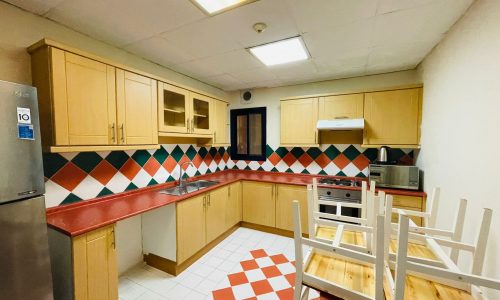 A brightly lit kitchen in a furnished apartment featuring wooden cabinets, a red countertop, patterned backsplash, and checkered floor tiles, with chairs inverted on a table to the right.