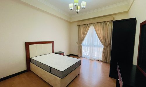 Interior of a bedroom in a furnished apartment with a large bed, cream walls, floor-length curtains, and a dark wardrobe.