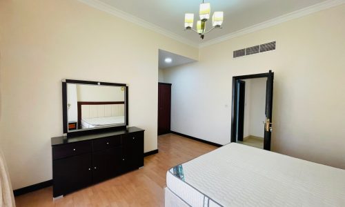 A spacious bedroom in a furnished apartment with a large bed, a dresser with a mirror, and two open doors leading to other rooms.