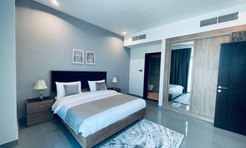 Modern bedroom with a large bed, white bedding, two bedside lamps, a gray rug, and an open door leading to another room.