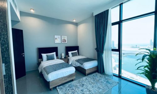 Modern bedroom with two twin beds, large window overlooking the sea, gray curtains, and minimalist decor.