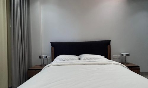 A modern bedroom in a 3 BDR apartment for rent, with a neatly made bed, flanked by two nightstands, under ambient lighting with grey curtains.