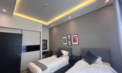 Modern fully furnished bedroom with two single beds, gray walls, ambient lighting, and framed artwork above a bedside table.