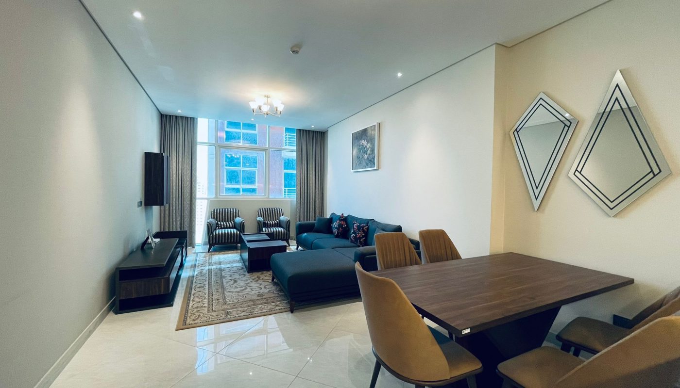 Luxury 1BR apartment in Juffair with a modern living room featuring a blue sofa, striped chairs, and a wooden dining table, lit by ceiling lights and adorned with decorative wall art.