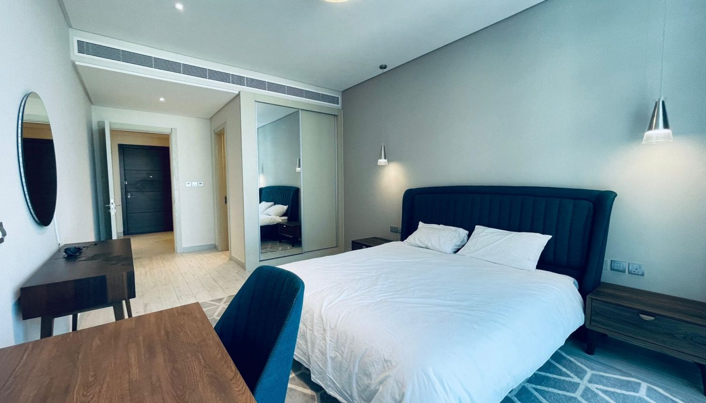 Luxury 1BR apartment bedroom with a large bed, overhead lighting, a desk, a chair, and an open door leading to another room.