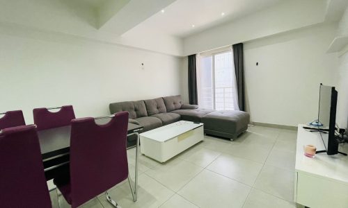 Modern living room in a luxury apartment with a grey sofa, white coffee table, flat-screen tv, and a dining area with a black table and purple chairs.