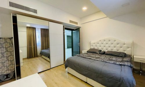 Modern apartment bedroom in Juffair with a large bed, mirrored wardrobe doors, and wooden flooring. Subtle lighting and a neutral color scheme complement the space.