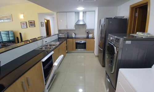 Modern kitchen interior in a flat in Amwaj with stainless steel appliances, wooden cabinets, and a narrow layout, featuring a double-door refrigerator and wall-mounted oven.