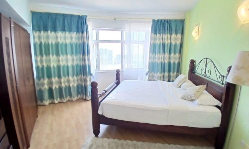 Bright bedroom in a modern flat featuring a large bed with white bedding, wooden headboard, patterned teal curtains, and adjacent wardrobe.