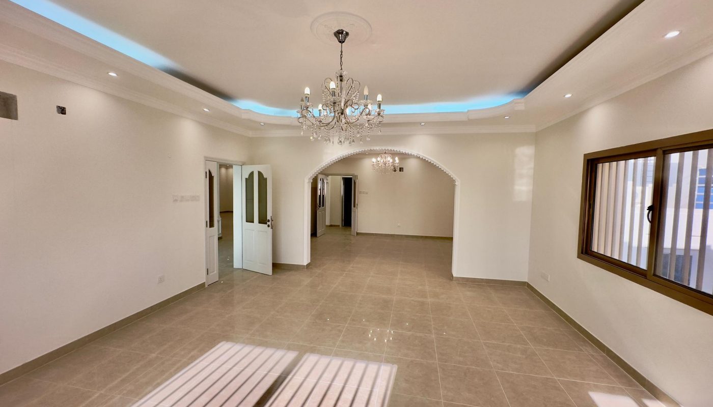 Spacious, empty living room with tiled floors, arched wall openings, recessed ceiling with blue led lights, and a crystal chandelier.