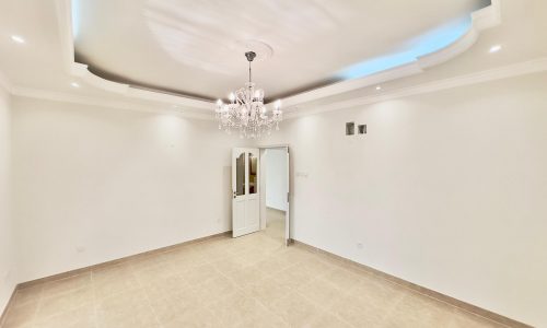 Spacious, empty room in a luxury apartment with white walls, coved ceiling with blue lighting, and a crystal chandelier. Tile flooring is visible and doors lead to other areas.