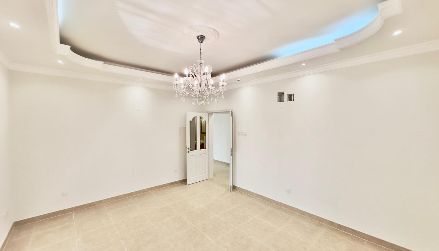 Spacious, empty room in a luxury apartment with white walls, coved ceiling with blue lighting, and a crystal chandelier. Tile flooring is visible and doors lead to other areas.