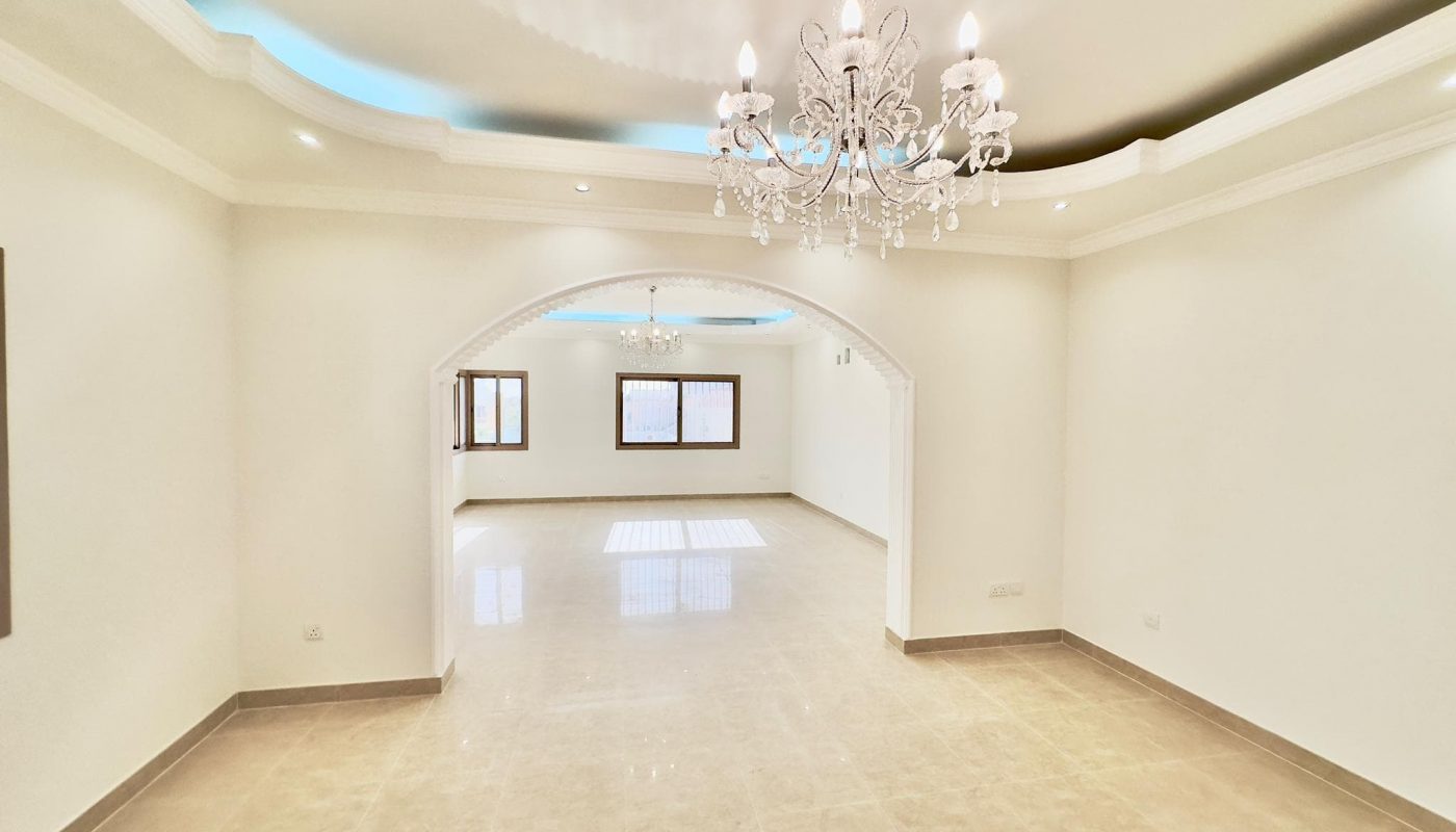Empty, spacious, brightly lit room with tiled floors, white walls, arched doorways, and a crystal chandelier.