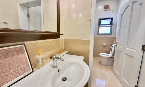 Modern bathroom interior in a luxury 3 bedrooms apartment, featuring a white sink, mirror, shower area, toilet, and beige tiles, with natural light from a small window.