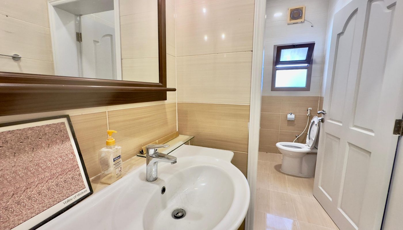 Modern bathroom interior in a luxury 3 bedrooms apartment, featuring a white sink, mirror, shower area, toilet, and beige tiles, with natural light from a small window.