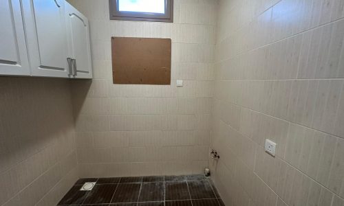 A small, partially tiled bathroom with a squat toilet, a hanging cabinet, and an empty board on the wall under a small window.