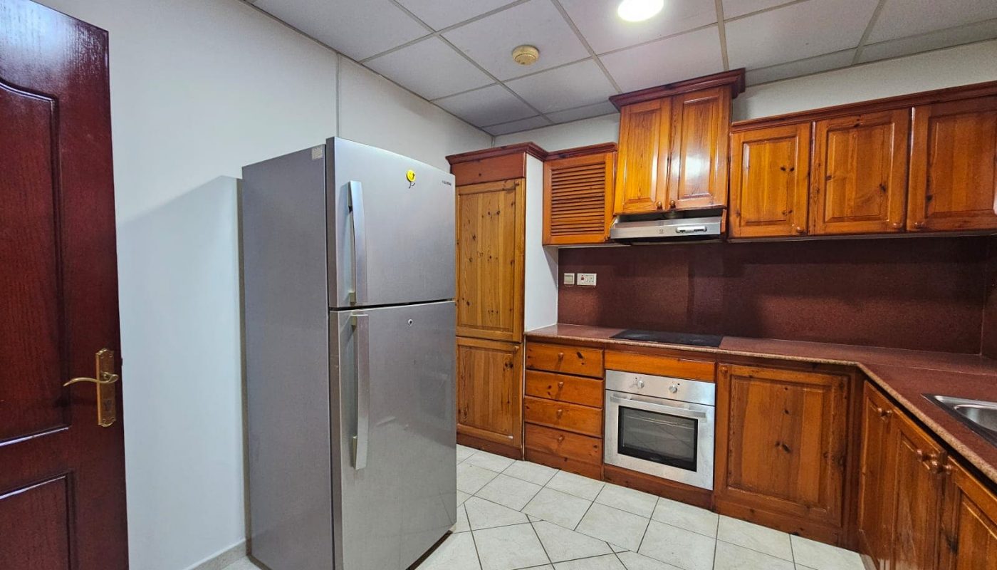 A kitchen in a flat for rent with wooden cabinets, a stainless steel refrigerator, built-in oven, and a microwave on a tiled floor.