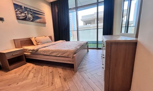 A modern bedroom in a luxury apartment with a wooden bed and floor, a side table, dresser, large windows with curtains, and a nautical-themed painting on the wall.