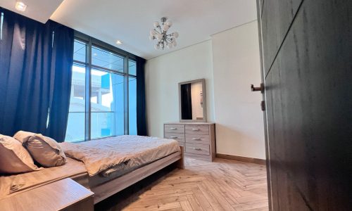 Modern bedroom in a luxury apartment, featuring a bed with gray bedding, wooden floor, chest of drawers, mirror, and large window, viewed from an open door.