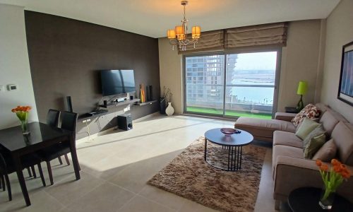 Modern living room with beige sofa, TV, dining area, and large window overlooking a water view in an Amwaj flat for sale. Bright and spacious interior design.