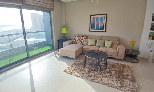 Modern living room in an Amwaj flat for sale, with beige sofa, chaise, and a glass coffee table on a shaggy rug. Large window with city view. Decor includes lamps, framed art, and flowers.