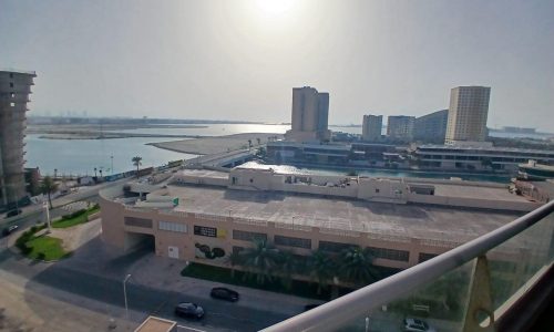 Wide-angle view from a balcony of a coastal Amwaj cityscape with buildings, a sunlit sea, and a clear sky.