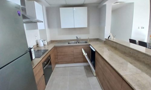 A modern flat for sale in Amwaj with wooden cabinets, granite countertops, and stainless steel appliances, including a fridge and microwave.