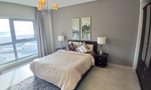 A modern bedroom in an Amwaj flat for sale, with a neatly made bed, two bedside tables with lamps, framed artwork above the bed, and a large window with a city view.