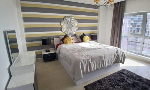 Modern bedroom in Amwaj with striped gray and yellow walls, a large bed with decorative pillows, mirrored nightstands, and a fluffy gray rug.