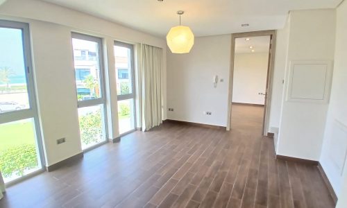 Bright, empty room in a modern villa for rent with large windows, wooden floor, and a modern light fixture, leading to another room with a view of greenery outside.