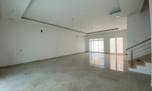 A spacious, empty room with white walls, a sloped ceiling, and large floor tiles in a villa for sale. There's natural light streaming through large windows and a glass door.