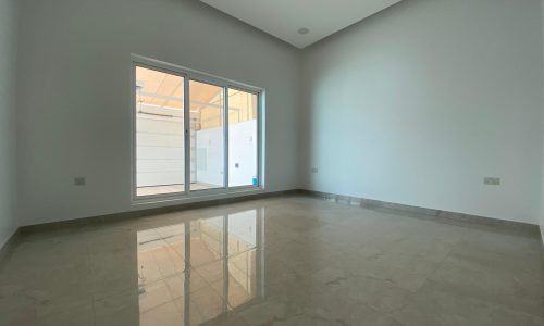 Empty room with polished marble floors, a large window, and a glass door leading outside, reflecting light on the surface of this Bani Jamra villa for sale.