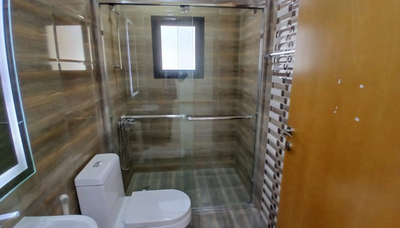 Modern bathroom with a glass-enclosed shower, toilet, towel rack, and a small window above the shower area. Walls and floor have a brown striped tile design.