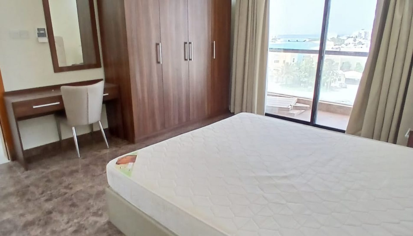 A bedroom with a double bed, wooden wardrobe, desk with a chair and mirror, large window with curtains, and marble floor.