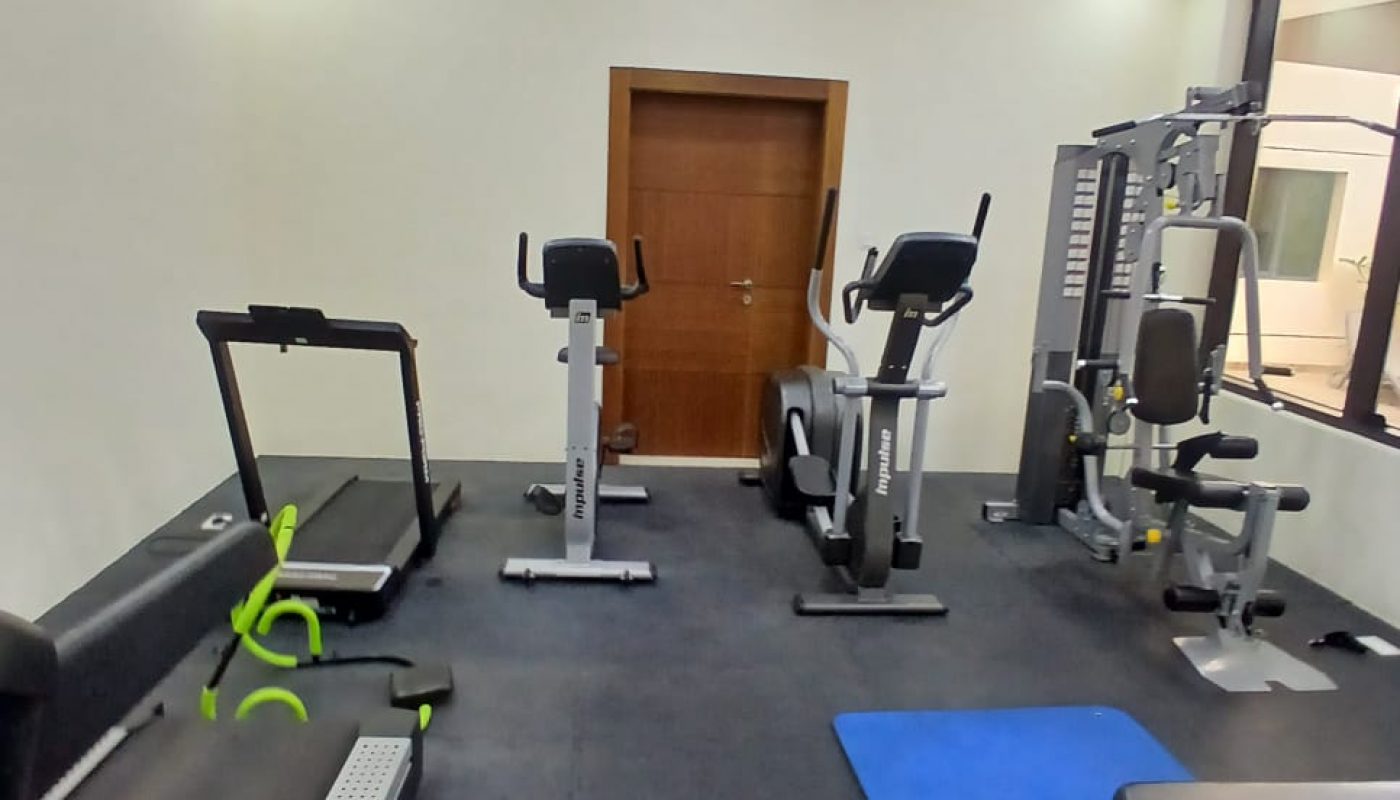A small gym room with treadmills, stationary bikes, a weight machine, a blue exercise mat, and a workout bench on a dark floor.