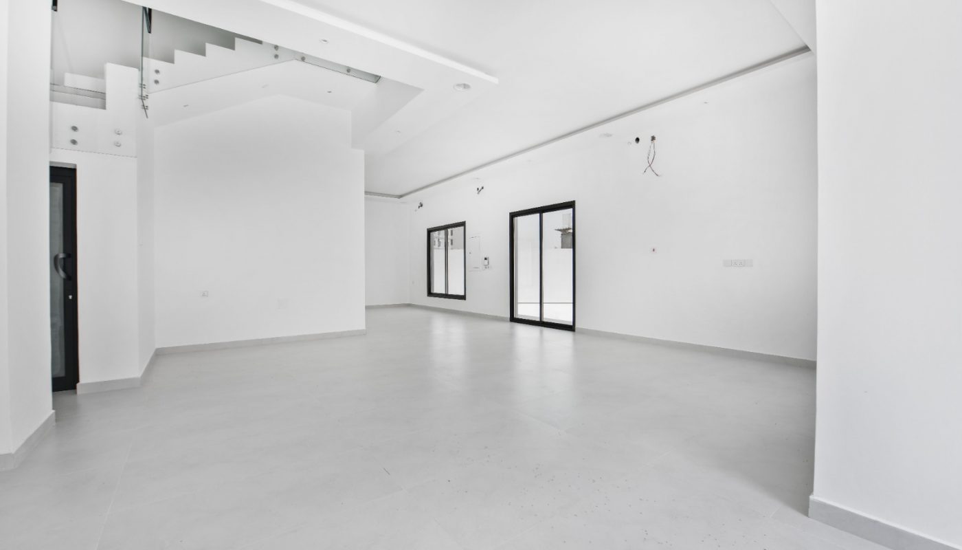 Spacious, empty modern room with white walls, large windows, tiled floor, recessed lighting, and a staircase in the background.