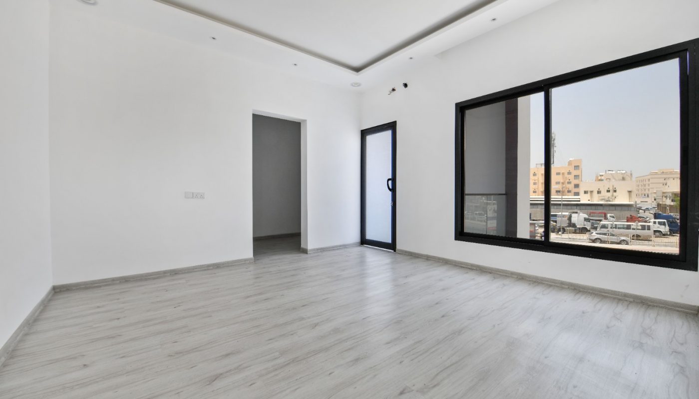 Empty, modern room with light gray wooden flooring, white walls, large window, and a glass door; view of buildings and parked vehicles outside.