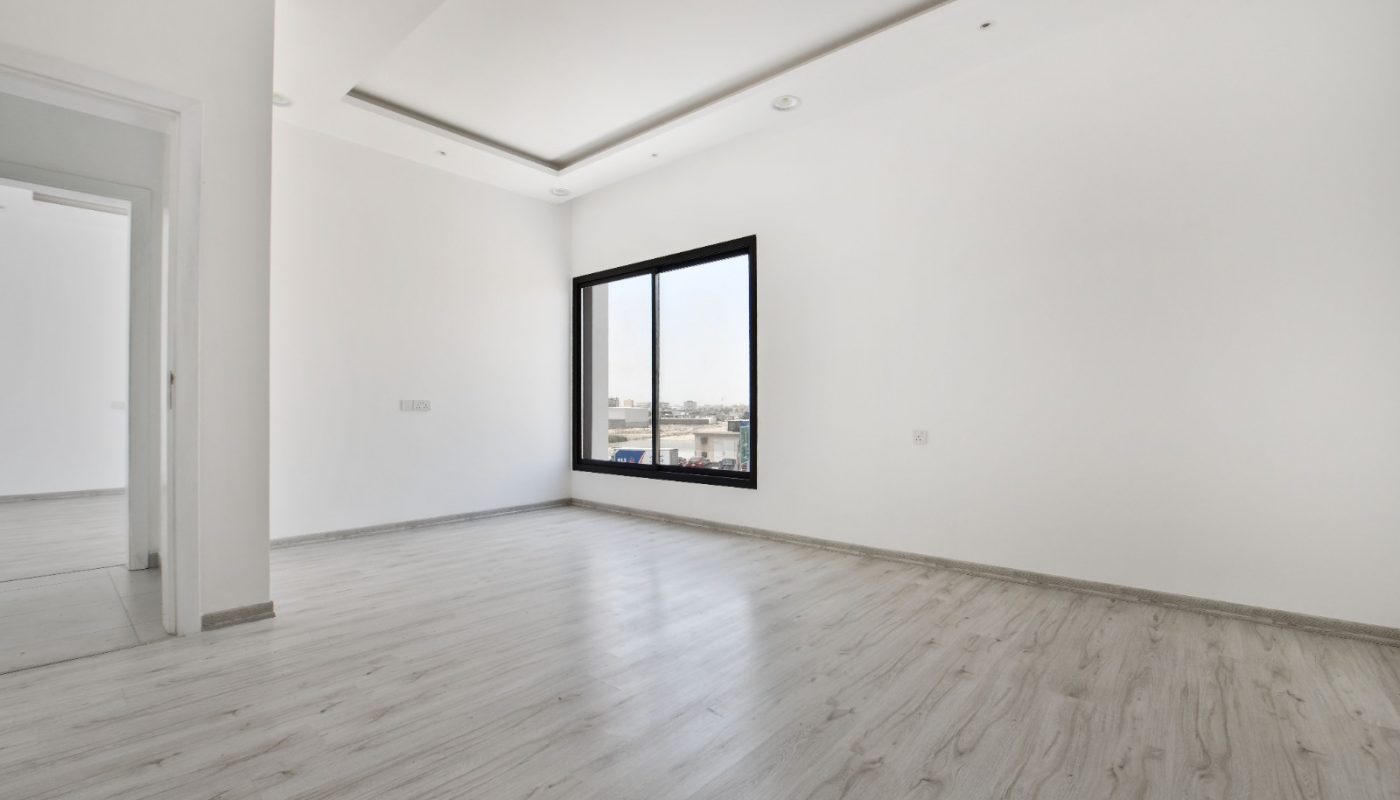 An empty room with white walls, light wood flooring, a large black-framed window, and a doorway leading to another space.