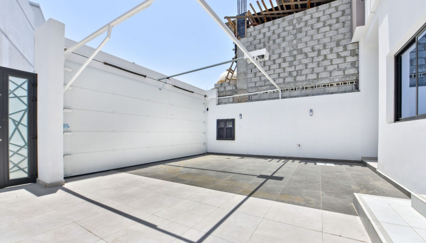 A modern, empty garage with a large open door and unfinished building structure in the background under clear daylight.