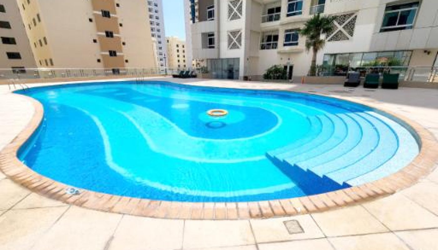 Outdoor swimming pool with blue tiles and curved steps, surrounded by apartment buildings and a patio area with seating.