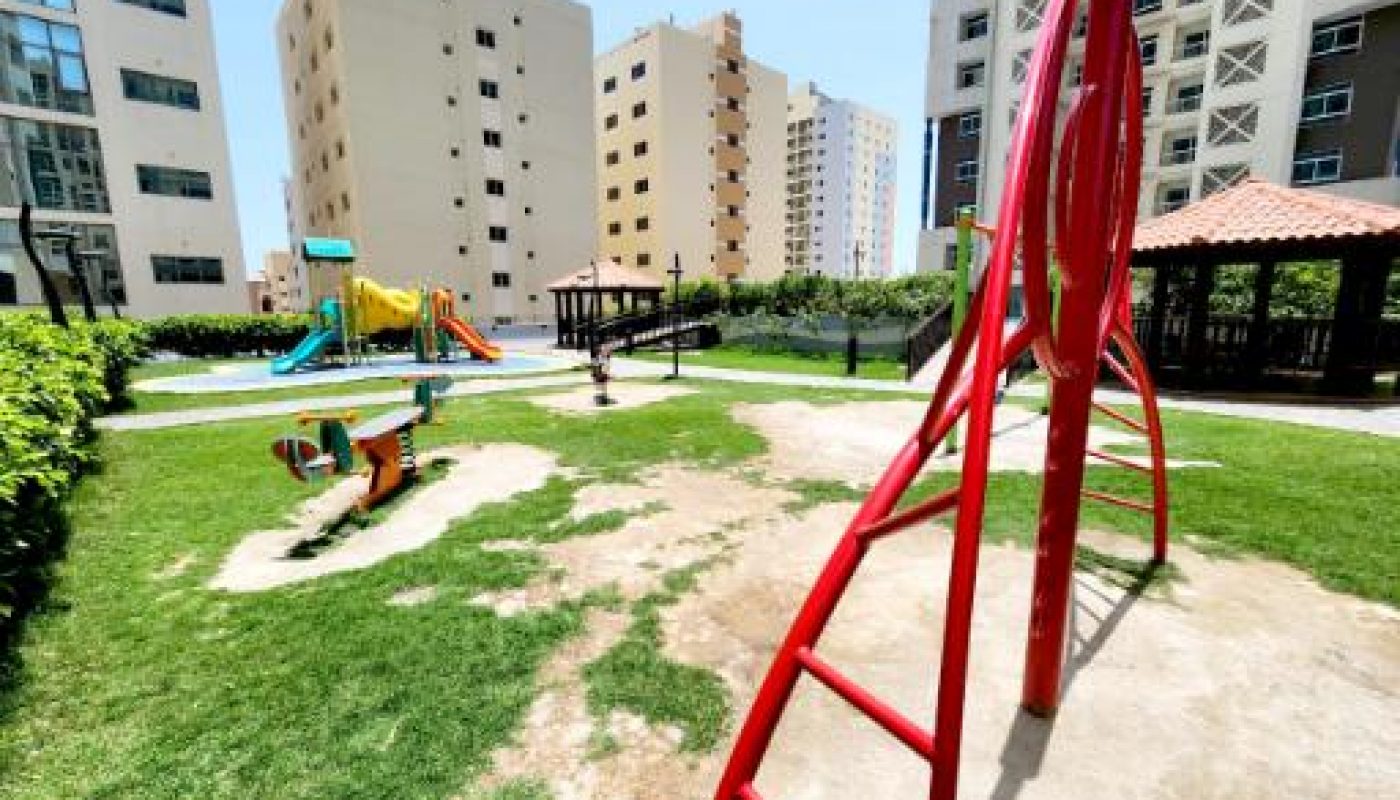 Playground with slides, climbing equipment, and seesaw surrounded by grass and sand, set among tall apartment buildings on a sunny day.