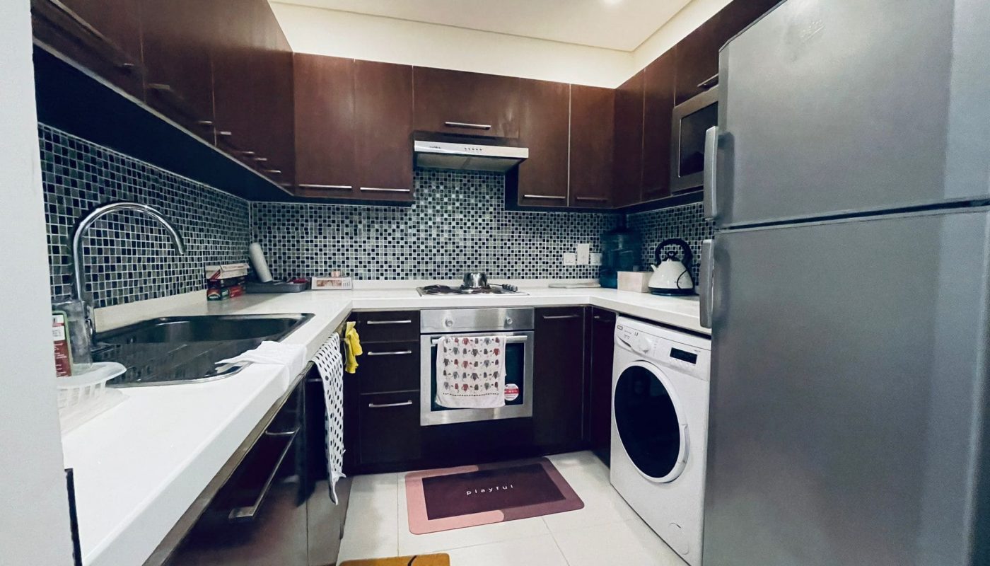 Modern kitchen with dark wood cabinets, stainless steel refrigerator, washing machine, sink, stove, and a tiled backsplash. A white countertop runs along the left and back walls. This luxury 1BR apartment in Juffair offers an elegant culinary space that seamlessly blends style and functionality.