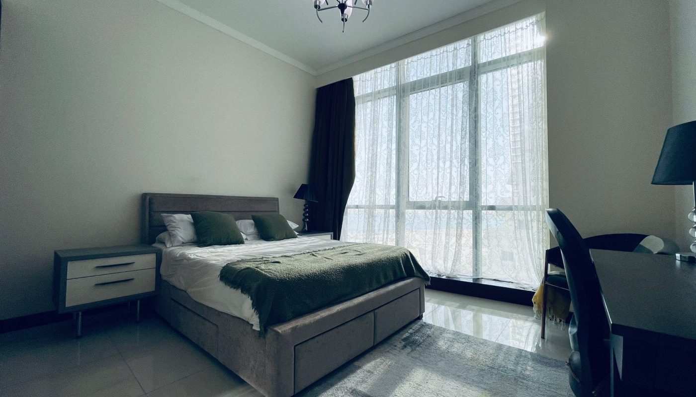 A well-lit bedroom in a luxury 1BR apartment in Juffair, featuring a large bed, two nightstands, a desk, a chair, and a modern chandelier. The room boasts tall windows with light curtains and a tiled floor with a patterned rug near the bed.