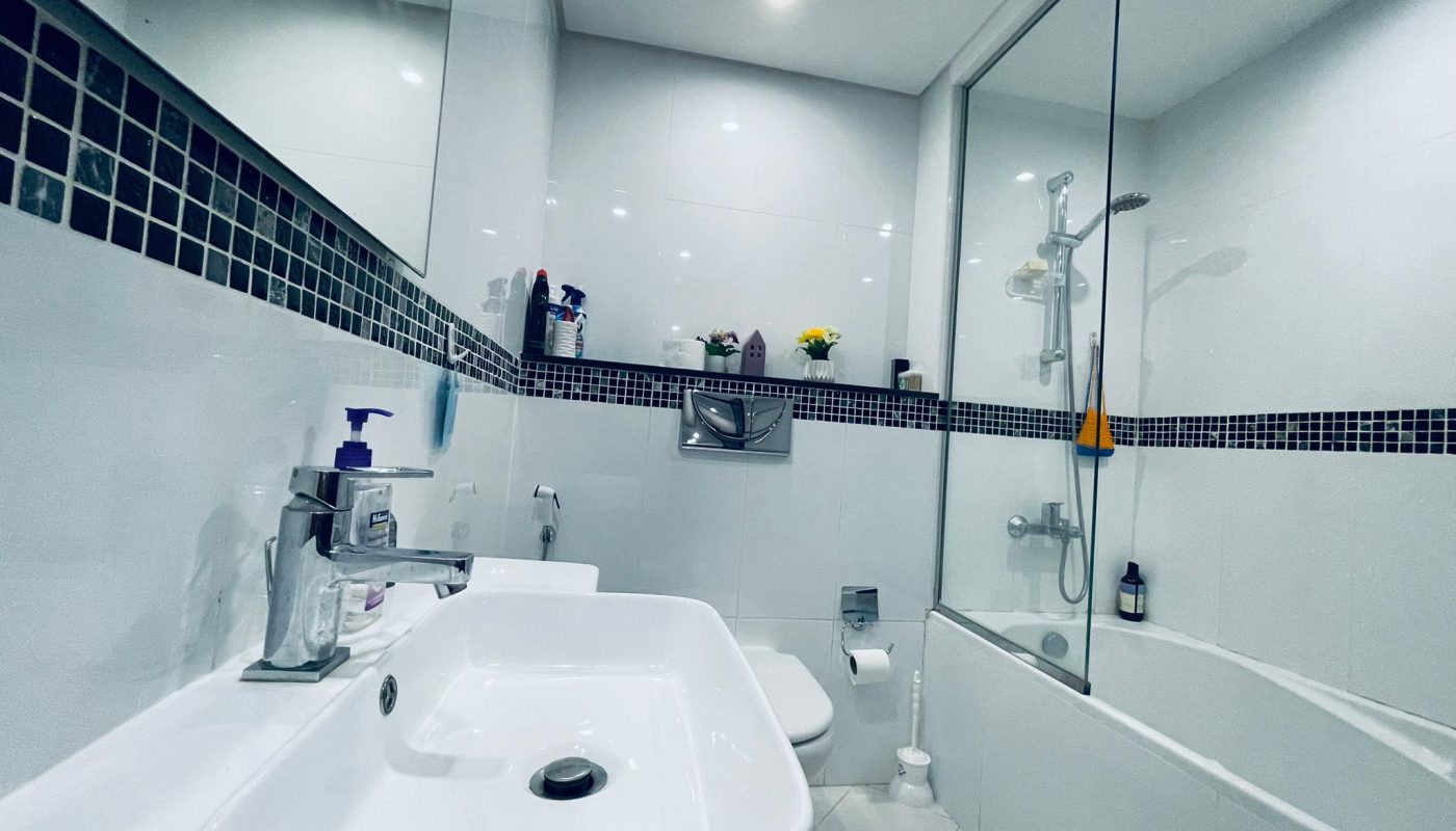 A modern bathroom in a luxury 1BR apartment in Juffair, featuring white tiles, a sink, toilet, and a bathtub with a glass shower enclosure. Shelves with toiletries line the wall above the toilet.