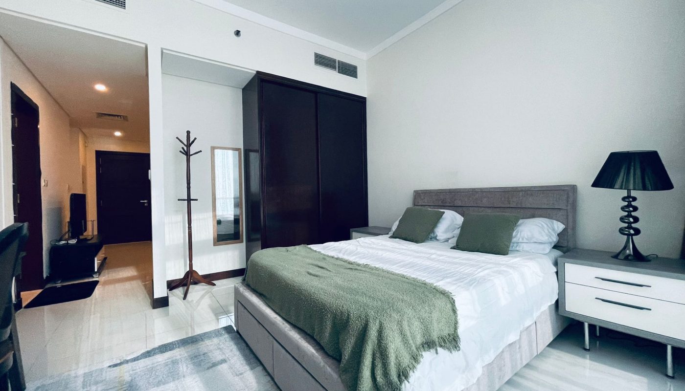 A luxury 1BR apartment in Juffair features a modern bedroom with a double bed, green cushions, and a matching throw. There is a nightstand with a lamp, a coatrack, a mirror, and a built-in wardrobe. The door and hallway are visible in the background.