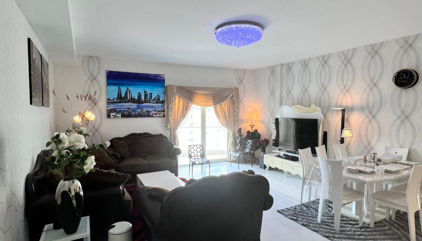 Bright living and dining area with brown sofas, a dining table set, large TV, wall art, and glass doors leading to a balcony.