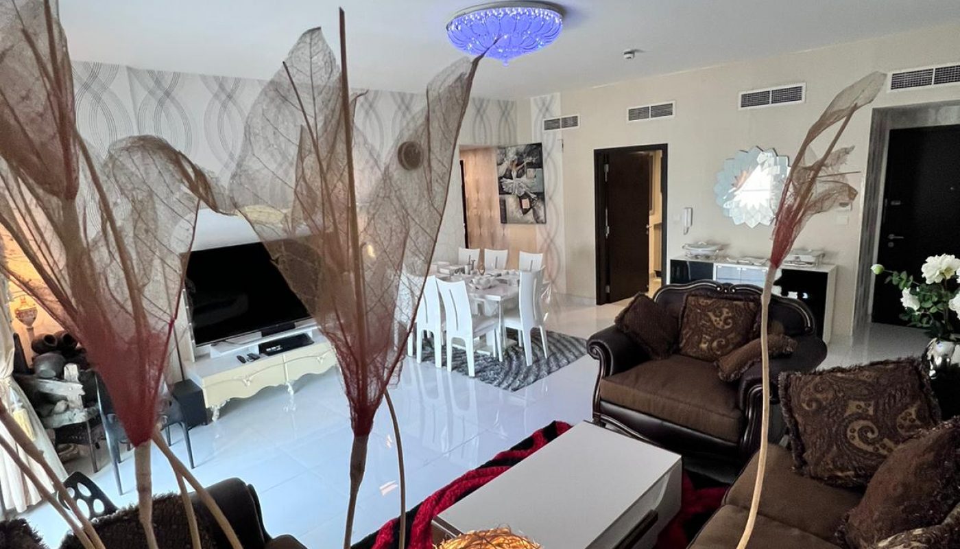 Modern living room with brown sofas, a white coffee table, decorative lights, and a TV. Dining area and entrance are visible in the background. Neutral tones and contemporary decor throughout.